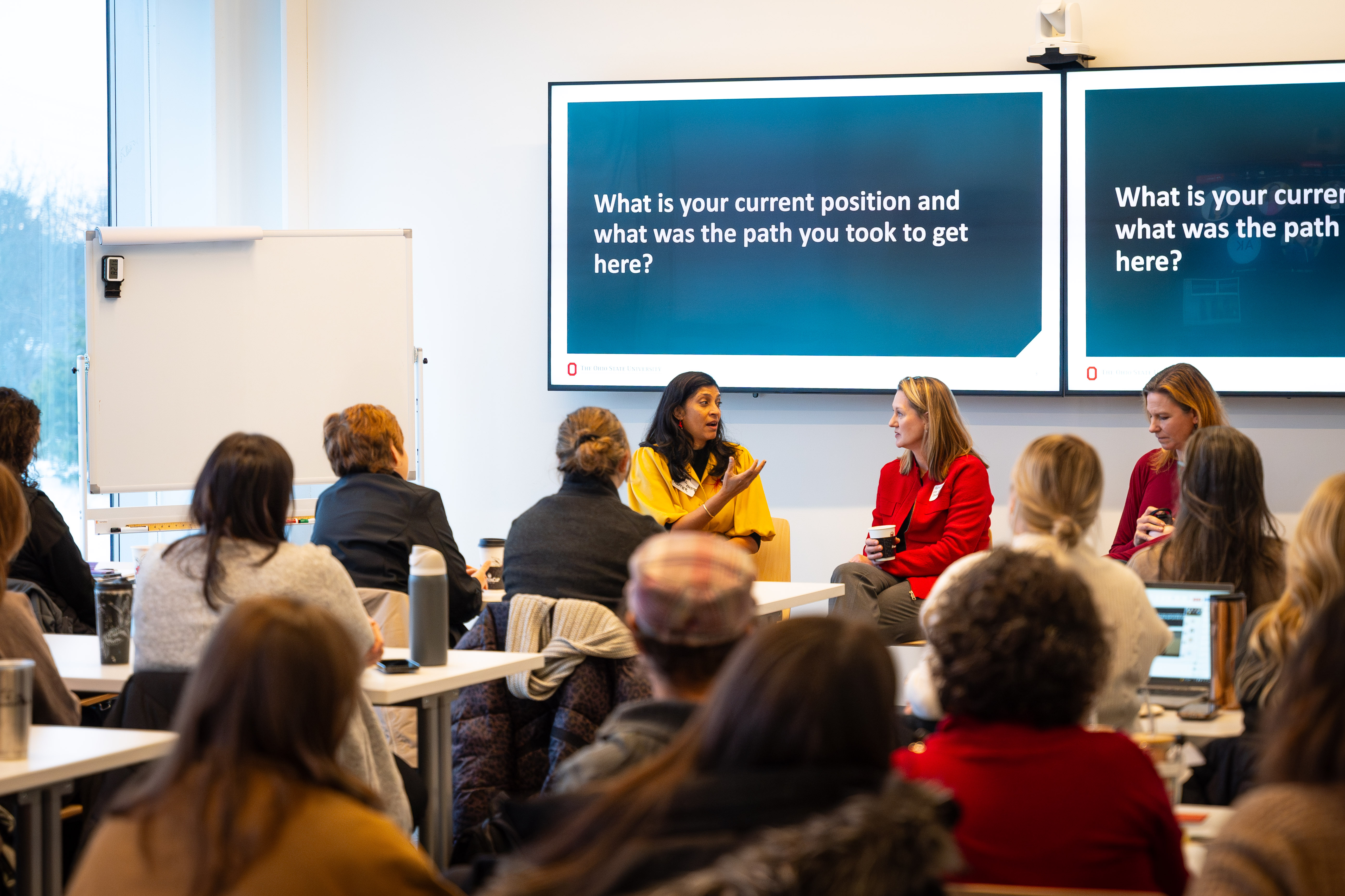 A group of women are seen sitting listening to three women speak who are seated at the front of a room.