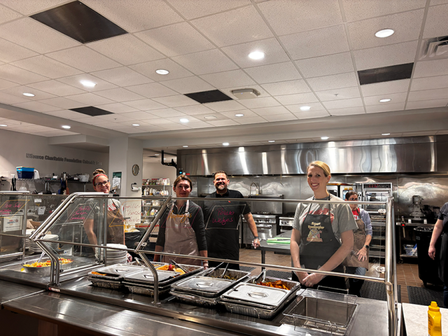 People stand behind a buffet serving table, smiling.
